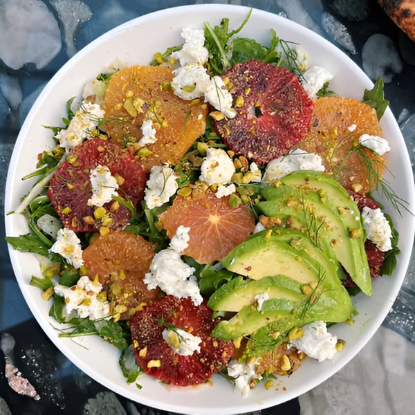A vibrant salad with blood oranges, avocado, goat cheese, pistachios, and greens in a white bowl.
