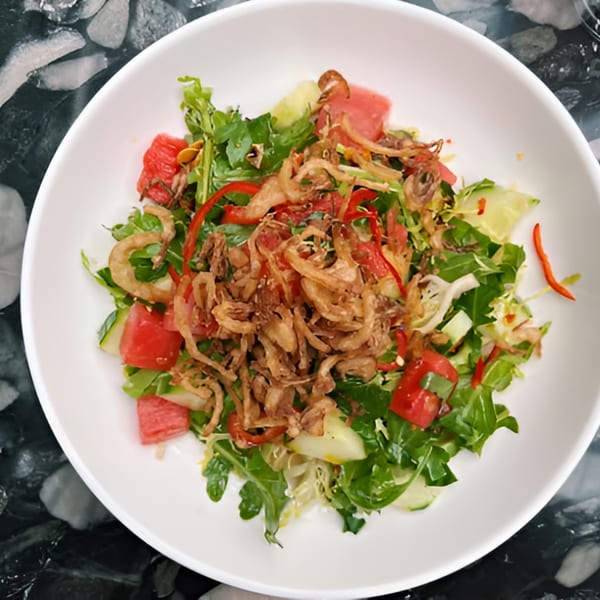 A vibrant salad with greens, watermelon, cucumber, red peppers, and crispy fried onions in a white bowl.