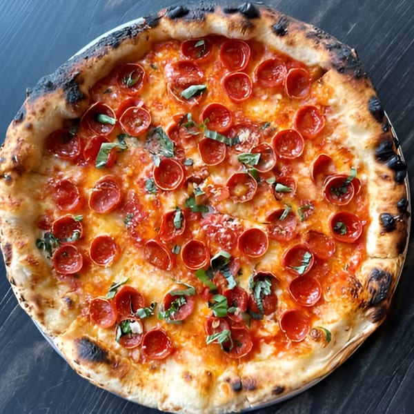A DAMNFiNE pepperoni pizza with fresh basil and a perfectly charred crust on a dark surface.